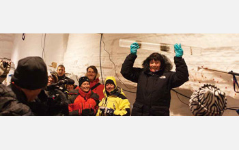 Photo of a woman in a snowsuit standing in a crowd inside a lab building holding a long ice core.