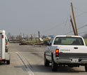 Photo of vehicles traveling down a road with downed telephone poles and collapsed houses.